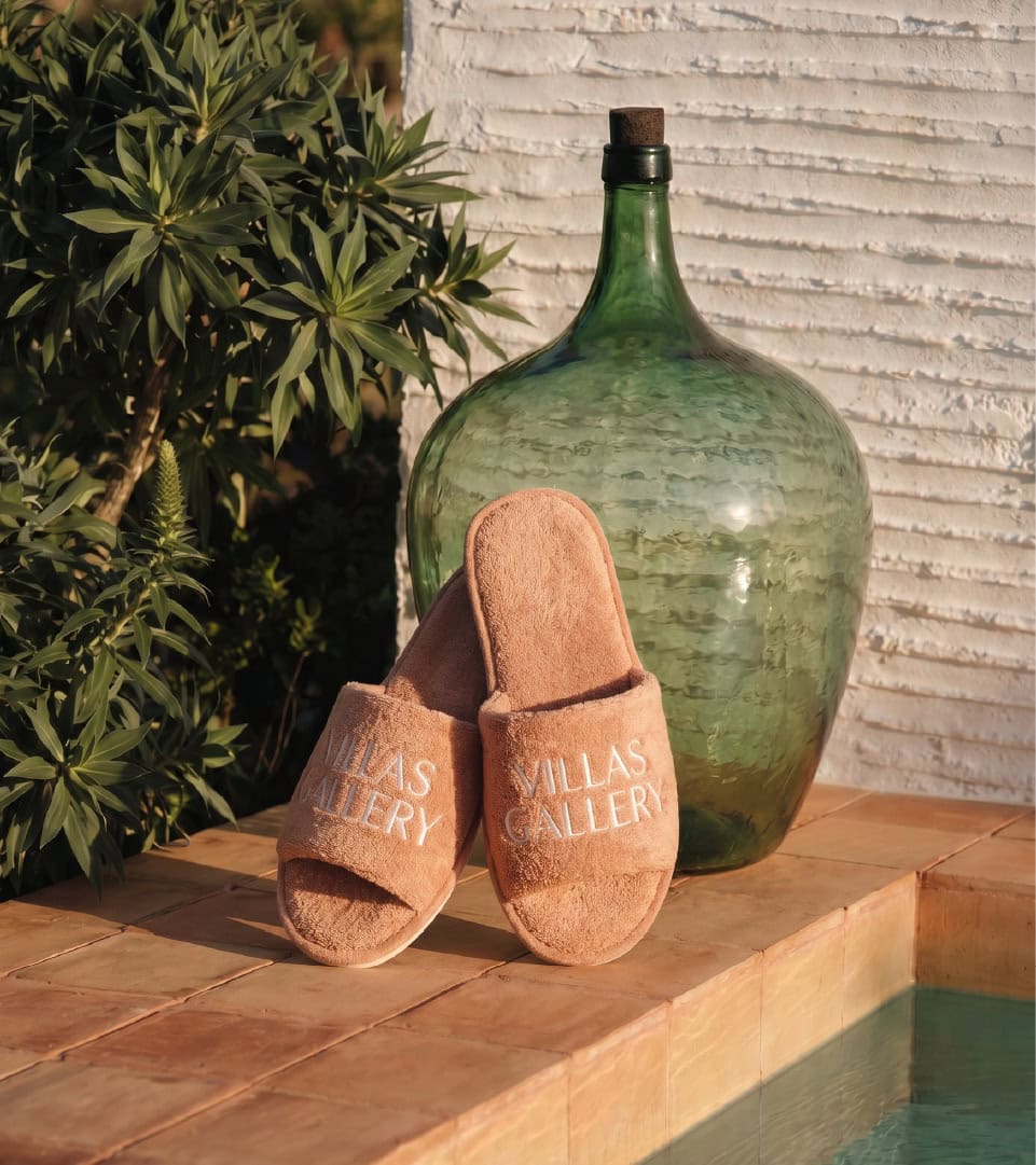 Zapatillas de casa con logo Villas Gallery junto a una botella de vidrio.