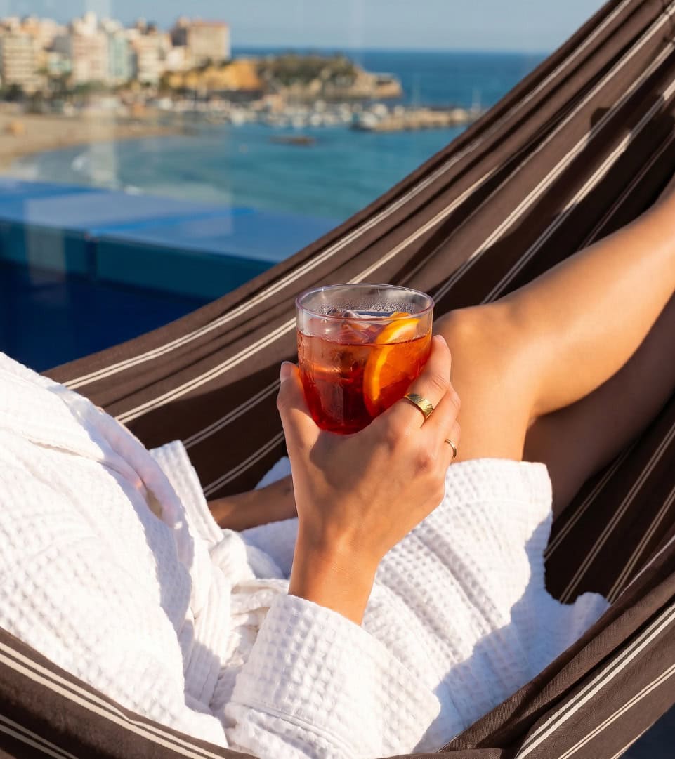 Persona disfrutando de una bebida en una hamaca con vista al mar.